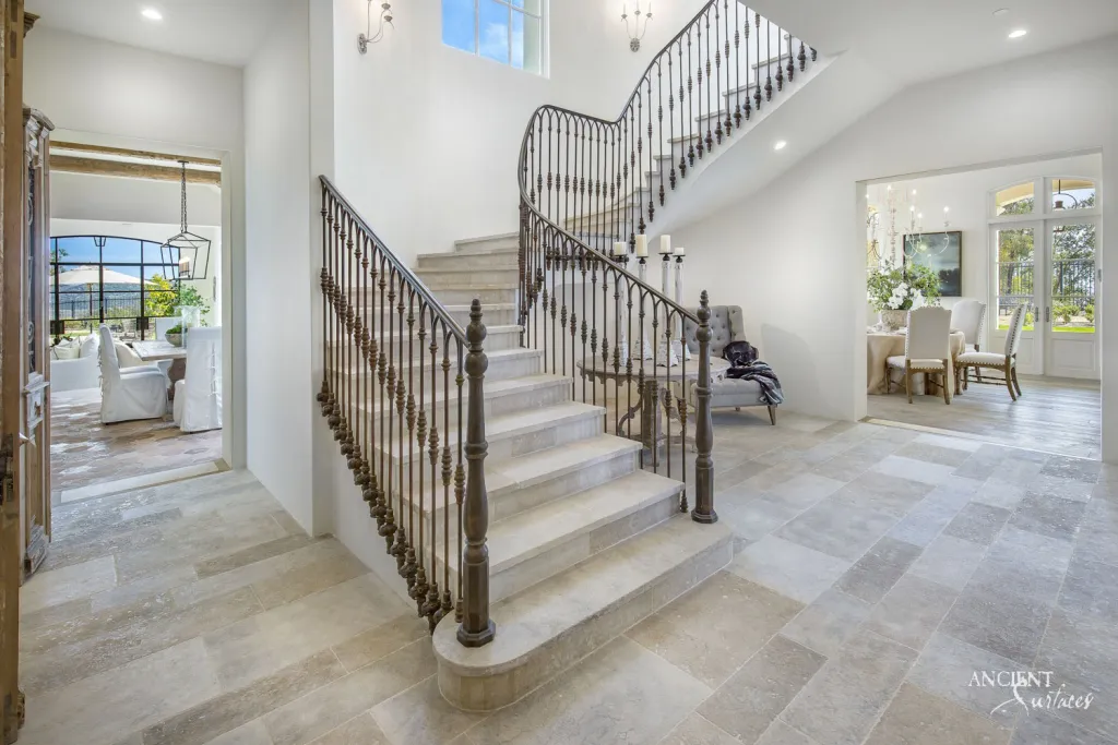 Curved limestone stair treads rising beside a wrought iron railing above a limestone paved foyer