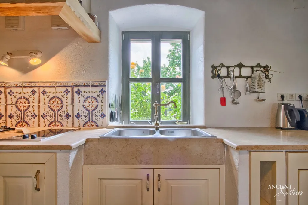 Pale stone kitchen counter and farmhouse sink beneath a hand painted tile backsplash