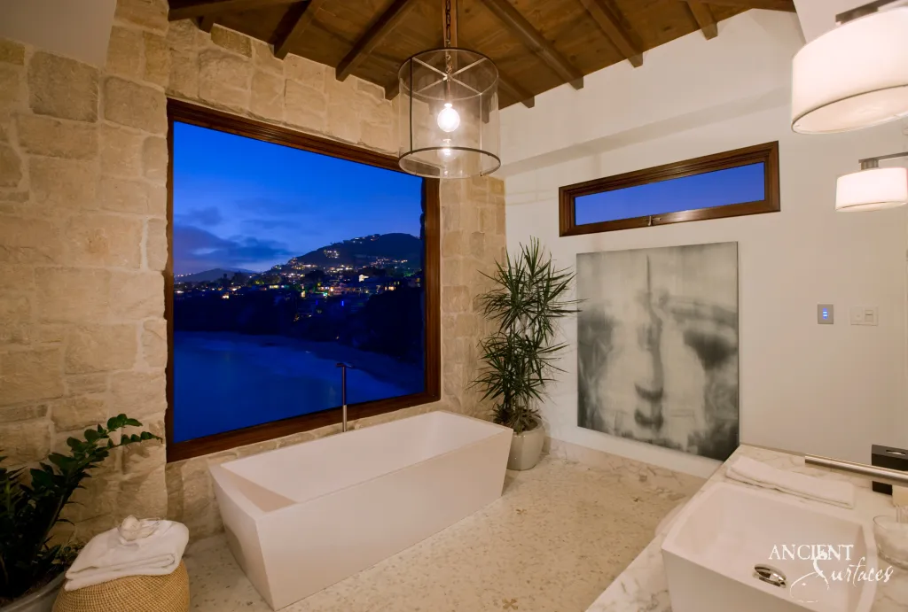 Freestanding tub beside limestone clad walls framing a nighttime ocean and hillside view