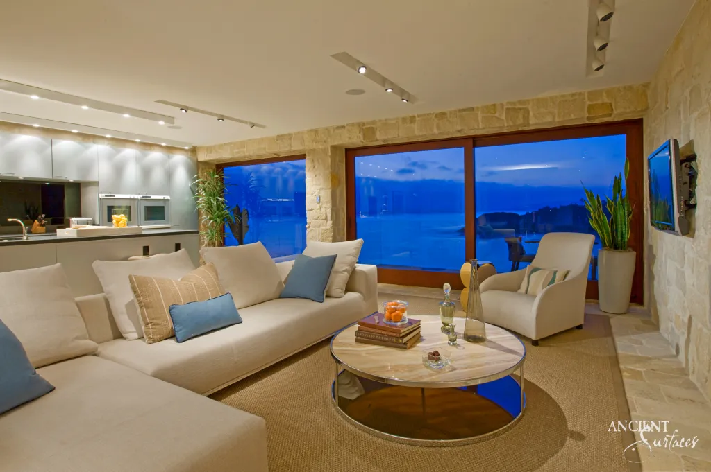 Sectional sofa facing floor to ceiling windows in a living room with limestone wall cladding, glowing at dusk over the sea